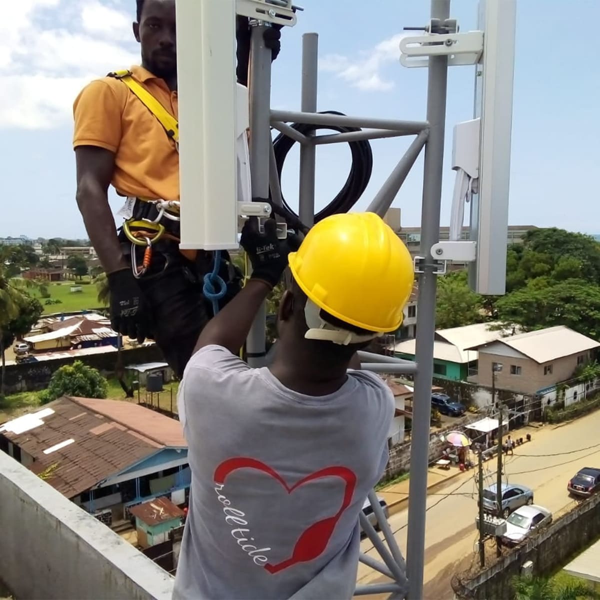 Deux ingénieurs, l'un portant un casque de chantier jaune et l'autre un harnais de sécurité, installant des antennes sectorielles sans fil sur un pylône surplombant Monrovia