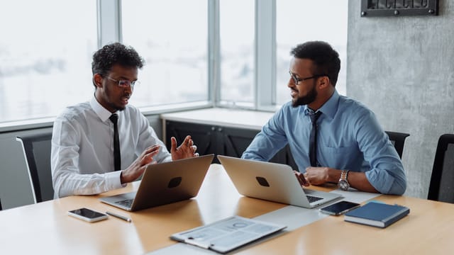 Two professionals in shirts and ties discussing a project over laptops at a meeting table