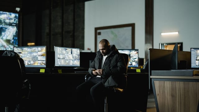 Security analyst at a monitoring desk with live camera and data feeds on several screens