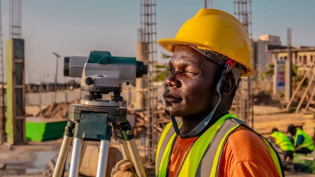 Surveyor in a hard hat and safety vest beside an optical level on a construction site