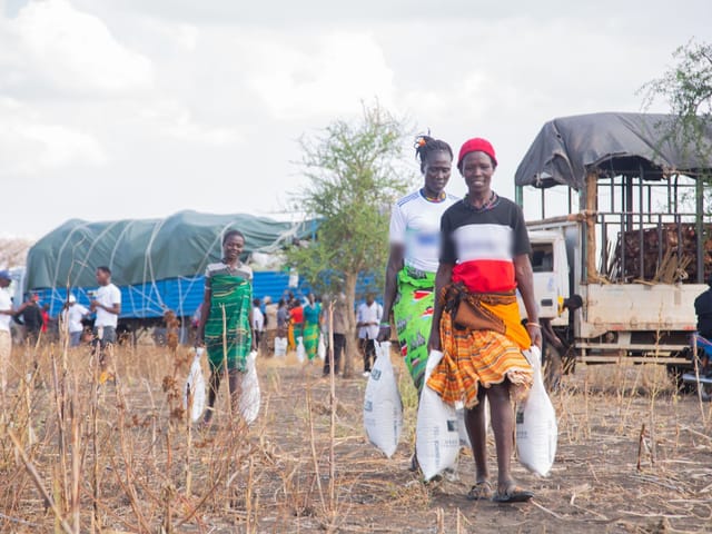 Women carrying bags of supplies from a distribution point, with trucks behind them